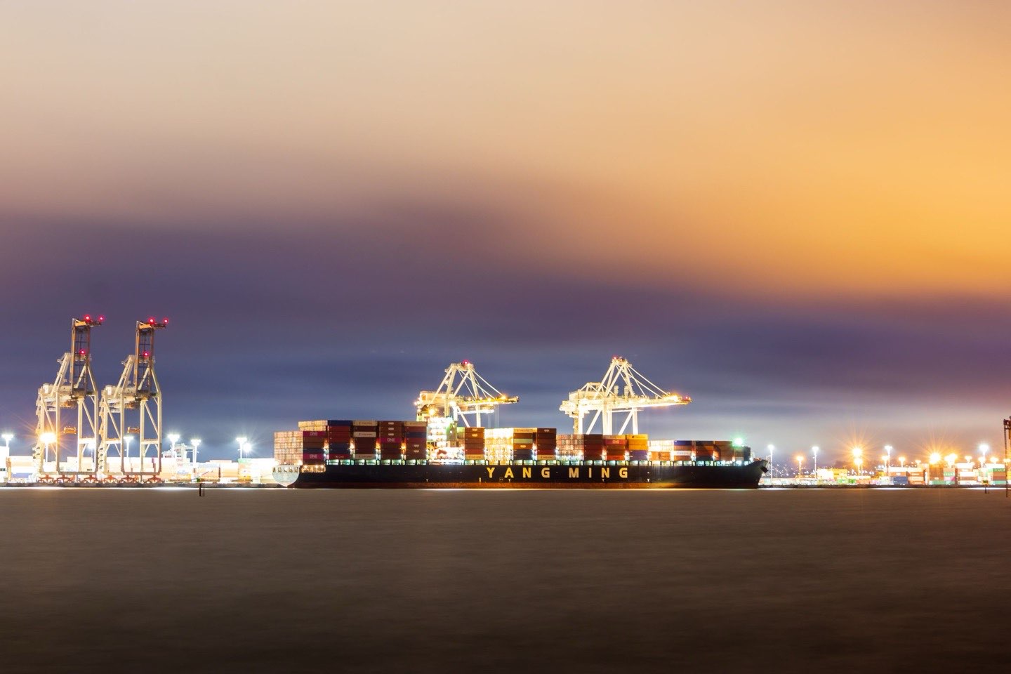 Container cranes and a docked ship at the Port of Oakland, lit against a dusk sky.